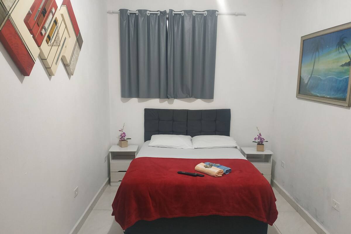 A bedroom with a bed, a red blanket, a remote control, and a painting on the wall.