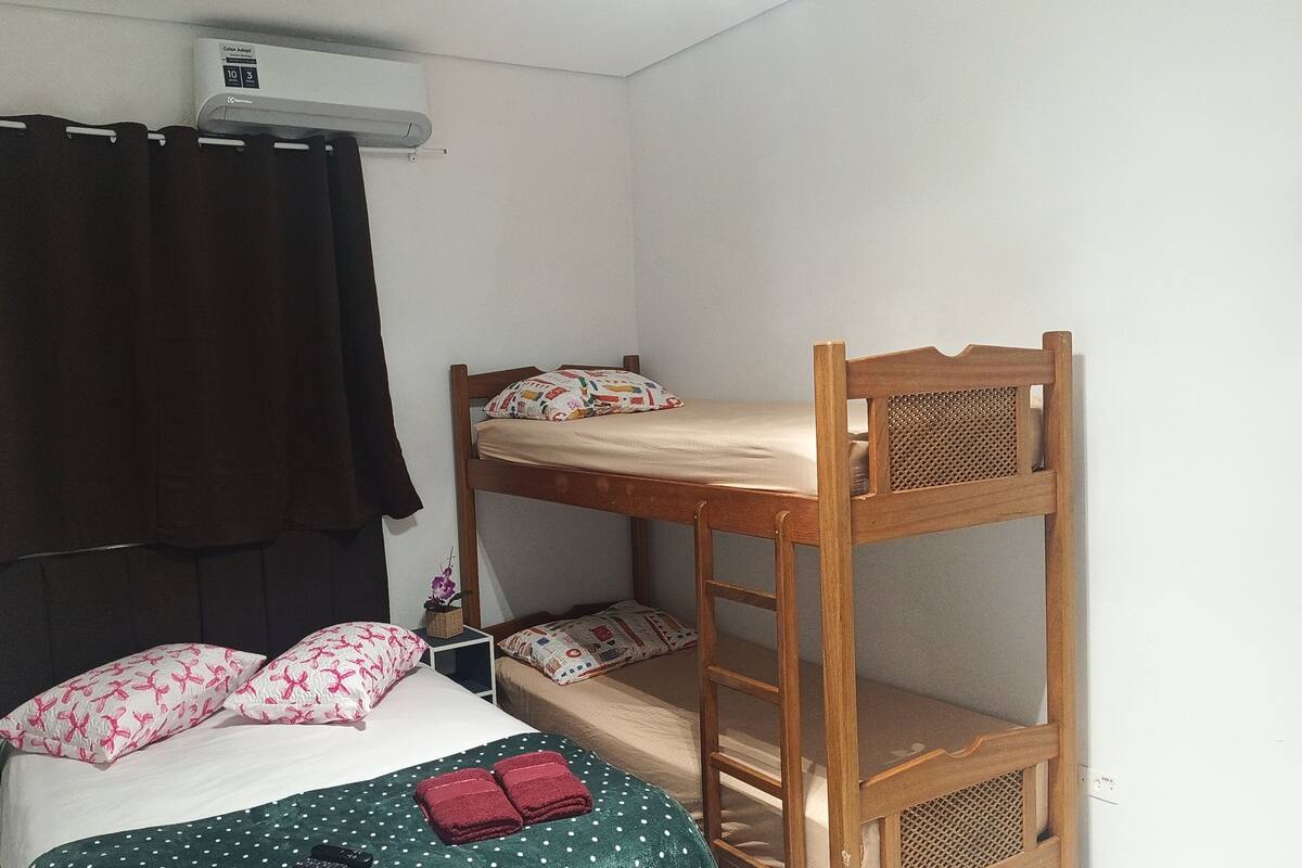 A bunk bed room with a single bed, a wooden bunk bed, a curtain, a wall-mounted air conditioner, and a small shelf with a plant.
