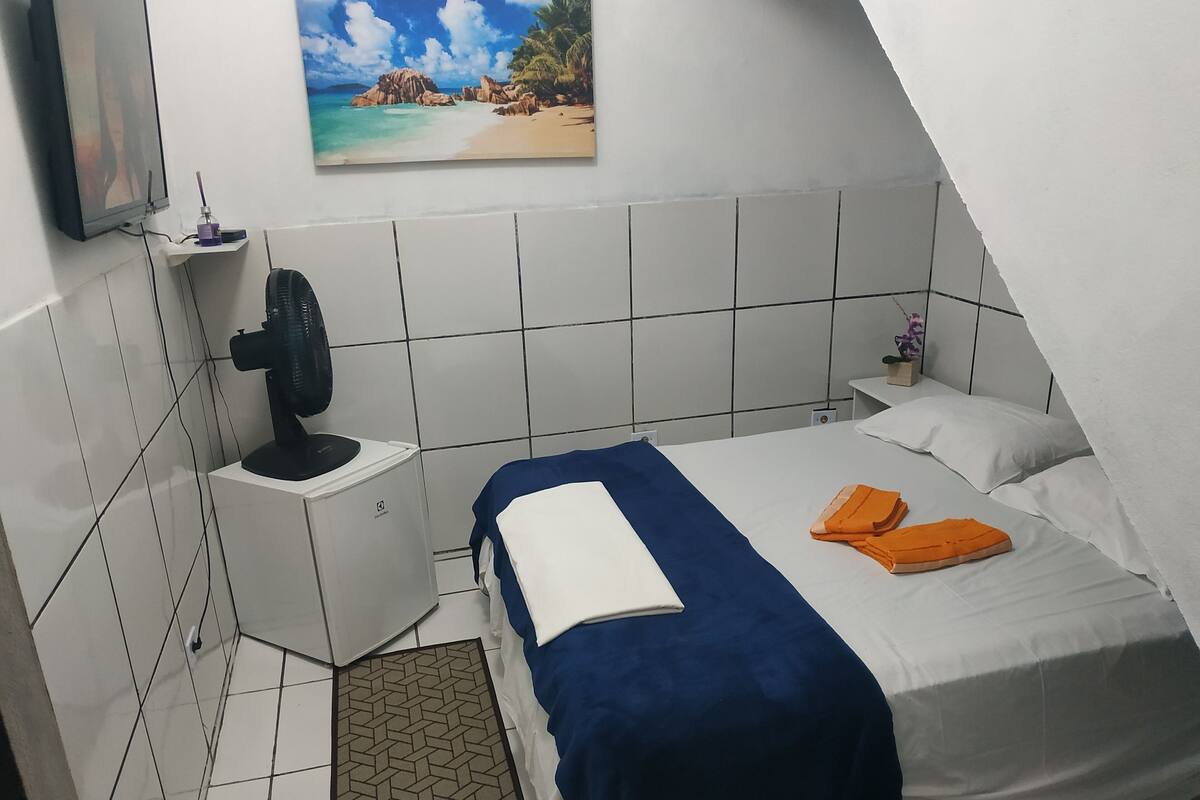 A bedroom with a bed, a nightstand, a fan, a painting of a beach, and a small plant.