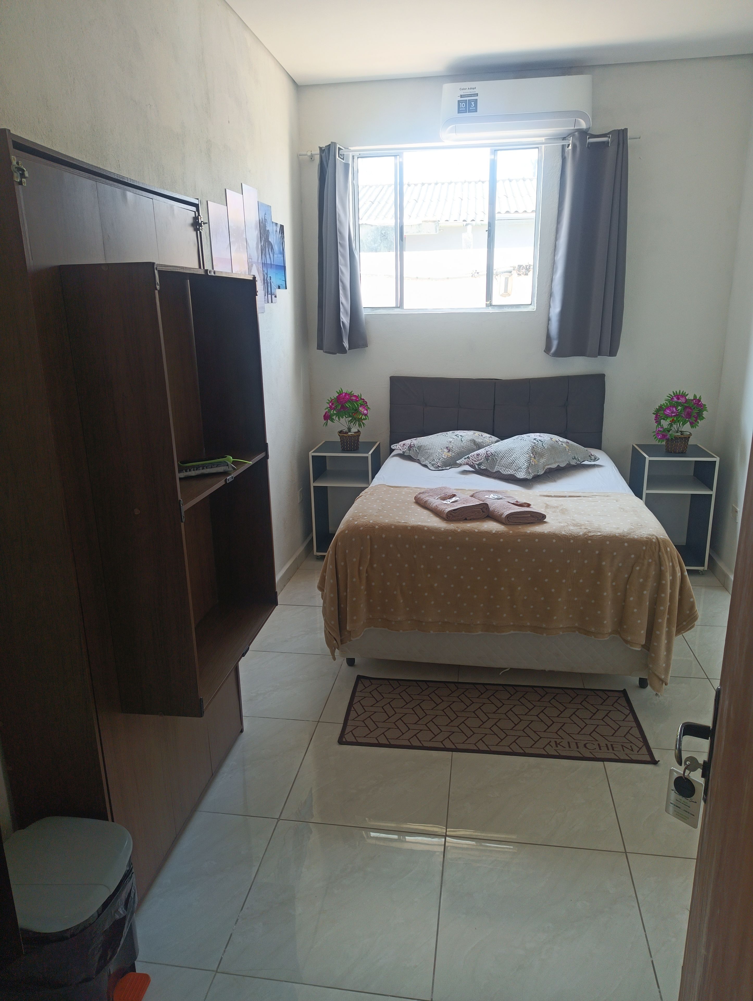 A bedroom with a bed, a wardrobe, two nightstands with flowers, a window with curtains, and a tiled floor.