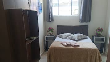 A bedroom with a bed, a wardrobe, two nightstands with flowers, a window with curtains, and a tiled floor.