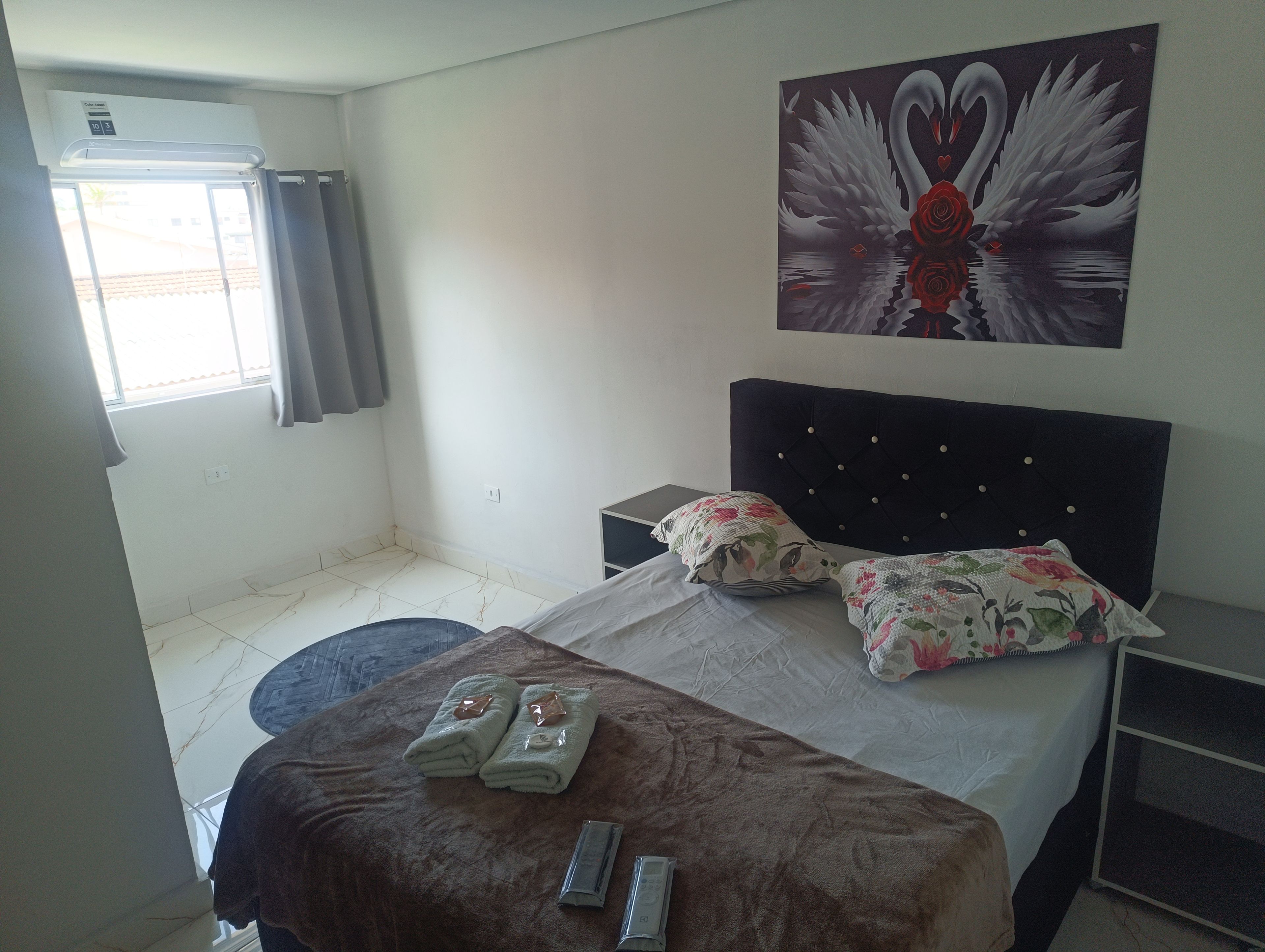 Ein Hotelzimmer mit Bett, Nachttisch, Wandklimaanlage, einem Fenster mit Vorhängen und einem Schwane-Gemälde.