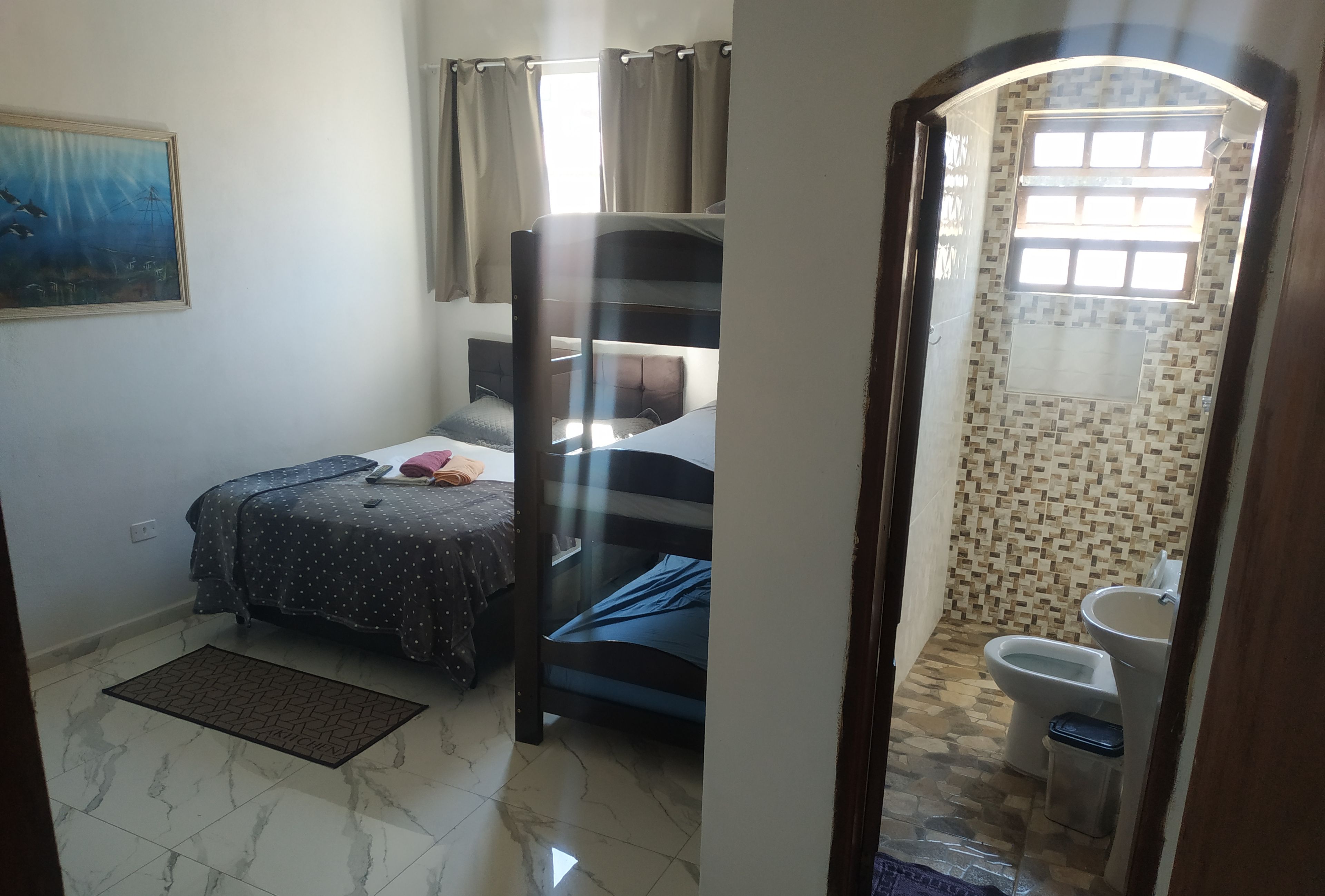 Una habitación con cama, ventana con cortinas, baño con lavamanos e inodoro, y suelo de baldosas.