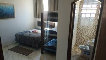 Una habitación con cama, ventana con cortinas, baño con lavamanos e inodoro, y suelo de baldosas.