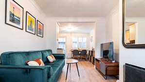 Un salon moderne avec un canapé de couleur turquoise, une petite table ronde et un téléviseur. Des tableaux encadrés ornent le mur, et un miroir est placé sur le côté droit.