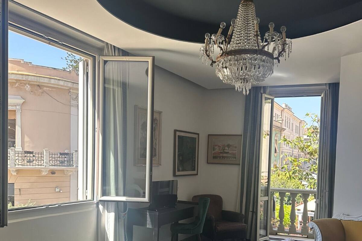 A hotel room with a large window, a desk, a chair, a bed with a green bedspread, and a chandelier.