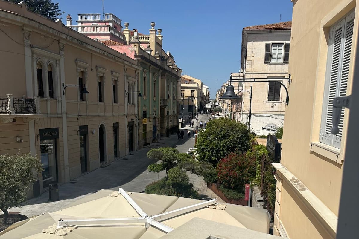 A narrow street with buildings on either side, a clear sky, and a balcony with a beige umbrella.