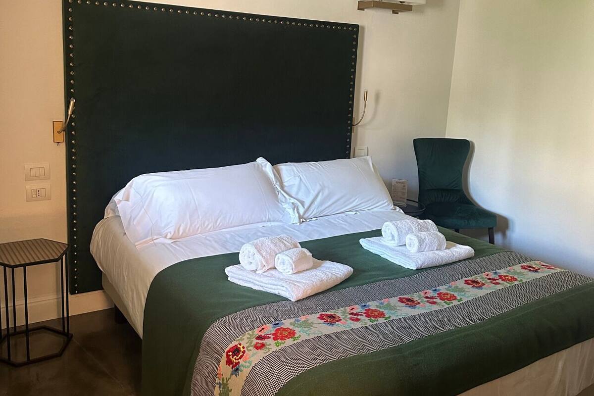 A neatly made bed with white linens and two folded towels, a dark green upholstered headboard, and a floral-patterned bedspread.