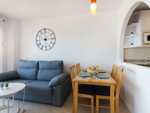 A living room with a grey sofa, a small round table, and a wooden dining table with chairs. A wall clock and a light fixture are visible.