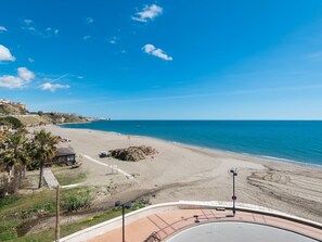A coastal road with a beach on the right, a clear blue sky, and a few buildings in the distance. - Front LINE Fuengirola Beach-Carvajal with terrace (FUENGIROLA)