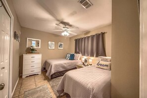 Quarto com duas camas, ventoinha no teto, cômoda e mesa de cabeceira com abajur.