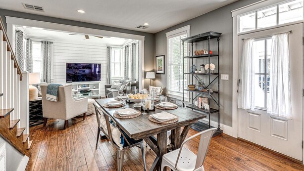 A spacious dining area with a wooden table set for four, surrounded by white chairs. A television is visible in the adjacent living room area, and a bookshelf filled with decorative items is against the wall. - Carlton Landing-Goose Bay (Carlton Landing)