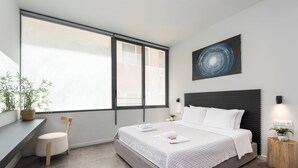 Un dormitorio moderno con un ventanal grande, una cama con ropa blanca, cabecera, mesita de noche con lámpara y un cuadro abstracto colgado en la pared.