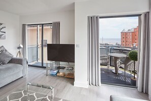 Un salon moderne avec un canapé, une table basse en verre et un téléviseur à écran plat. Il y a un balcon avec vue sur la ville et un immeuble.