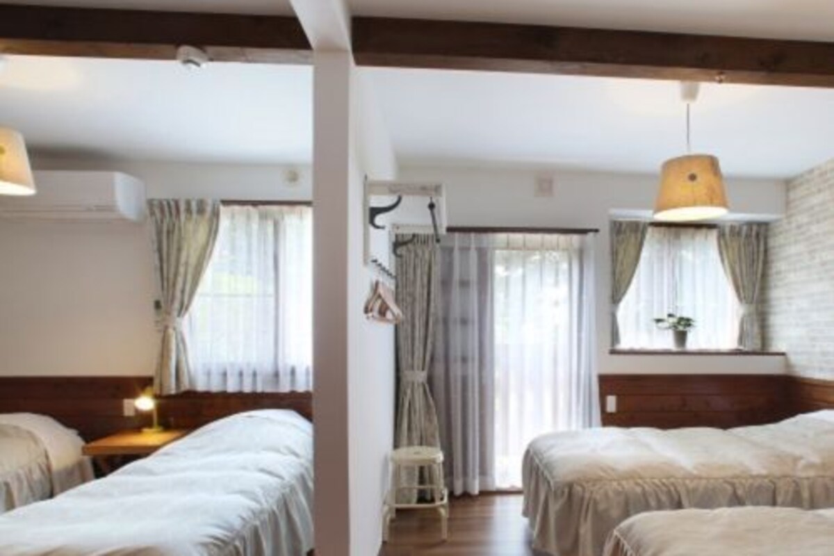 A room with two beds, wooden floors, a window with curtains, and a hanging light fixture.