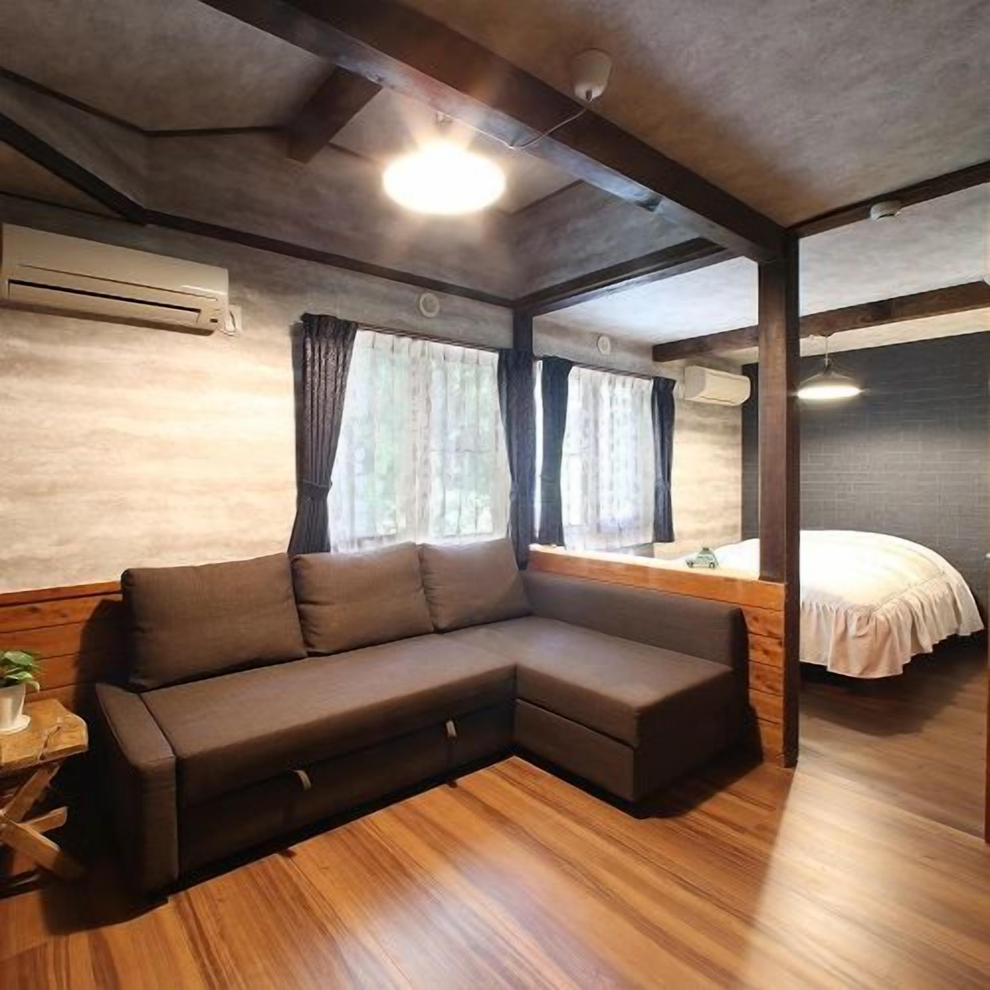 A room with a sectional sofa, a bed, wooden flooring, and a window with curtains.