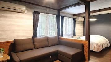 A room with a sectional sofa, a bed, wooden flooring, and a window with curtains.