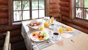 Una mesa de desayuno con diversos alimentos, entre ellos huevos, tostadas y fruta, ubicada en una cabaña con paredes de madera y ventanas amplias.