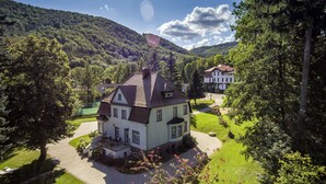 Un grand bâtiment blanc, surmonté d’un toit sombre, entouré d’une végétation luxuriante et situé dans un paysage vallonné.