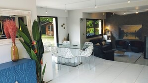 A modern living room with a glass dining table, white chairs, a black sofa, and a coffee table. There is a large window with a view of greenery outside.