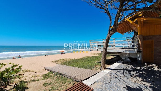 A beach view with a wooden deck, a beach umbrella, and a building with a thatched roof. - Flat pé na areia com vista internet e ar (Macaé)