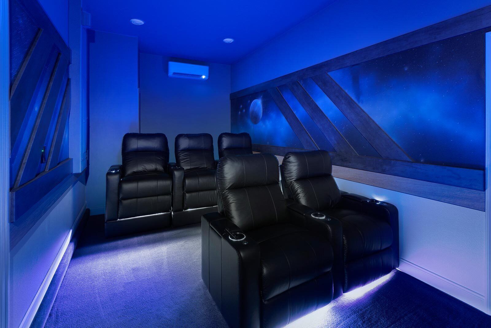 Sala de cine en casa con cuatro sillones de cuero negro, un proyector instalado en el techo y un mural en la pared que representa un cielo estrellado.