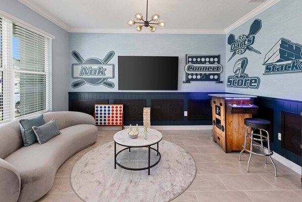 A modern living room with a sofa, a coffee table, and a TV mounted on the wall. There are wall decorations, a dartboard, and a bar area with stools. - Mermaid Magic | New Beach Home w Sea Themed BR, Pool, Theater, Arcade, Game Rm (Melbourne Beach)