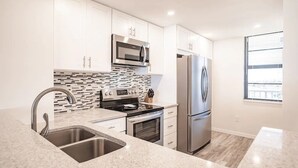 A modern kitchen with white cabinets, stainless steel appliances, a tiled backsplash, and a window providing natural light. - Beachfront I Full Coastal Contemporary Renovation (Marco Island)