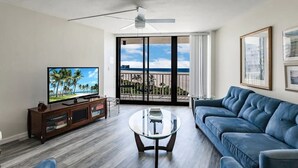 A living room with a television, a blue sofa, a glass coffee table, a ceiling fan, and a balcony with a view of the ocean. - Beachfront I Full Coastal Contemporary Renovation (Marco Island)