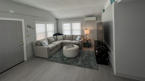 A living room with a sectional sofa, ottoman, and a flat-screen TV. - Walk to the Beach! Two bedroom cottage in Swifts Beach community. (Wareham)