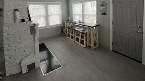 A room with a white brick accent wall, a dining table with benches, two windows with blinds, and a door. - Walk to the Beach! Two bedroom cottage in Swifts Beach community. (Wareham)