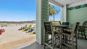 Een overdekt terras met strandstoelen en een tafel, met uitzicht op een zanderig strand met kleurrijke strandstoelen en een palmpijlboom.