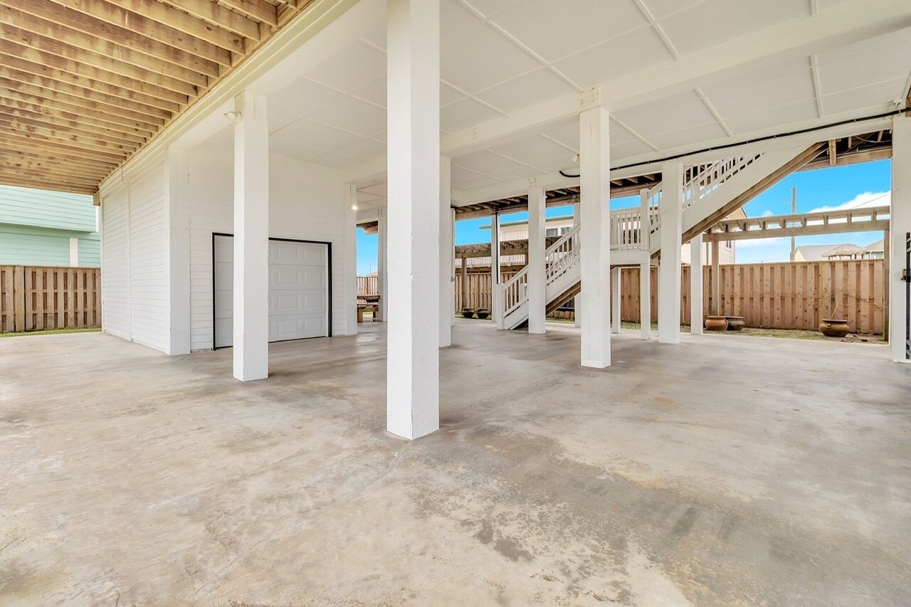 A spacious covered patio area with concrete flooring and white pillars, leading to a wooden deck and a staircase.