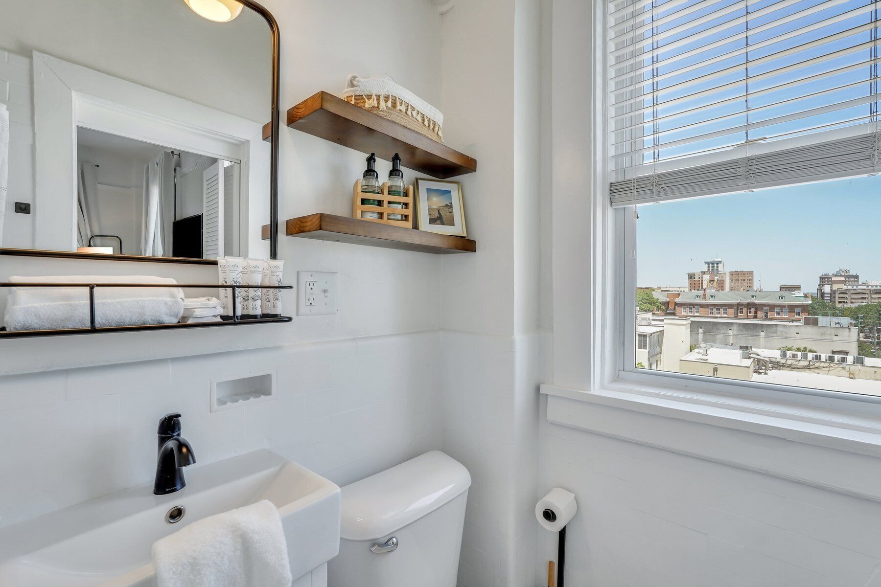 A modern bathroom with a white sink, black faucet, and a toilet. There are wooden shelves above the toilet holding toiletries and a framed picture. A window with blinds provides a view of buildings outside.