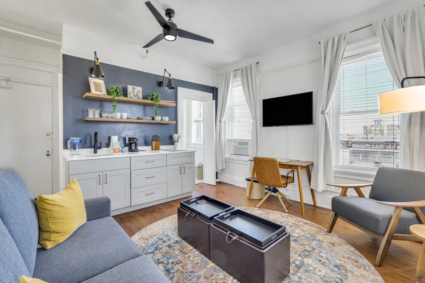A modern living room with a sofa, coffee table, and a TV. There is a kitchen area with shelves and a dining table.