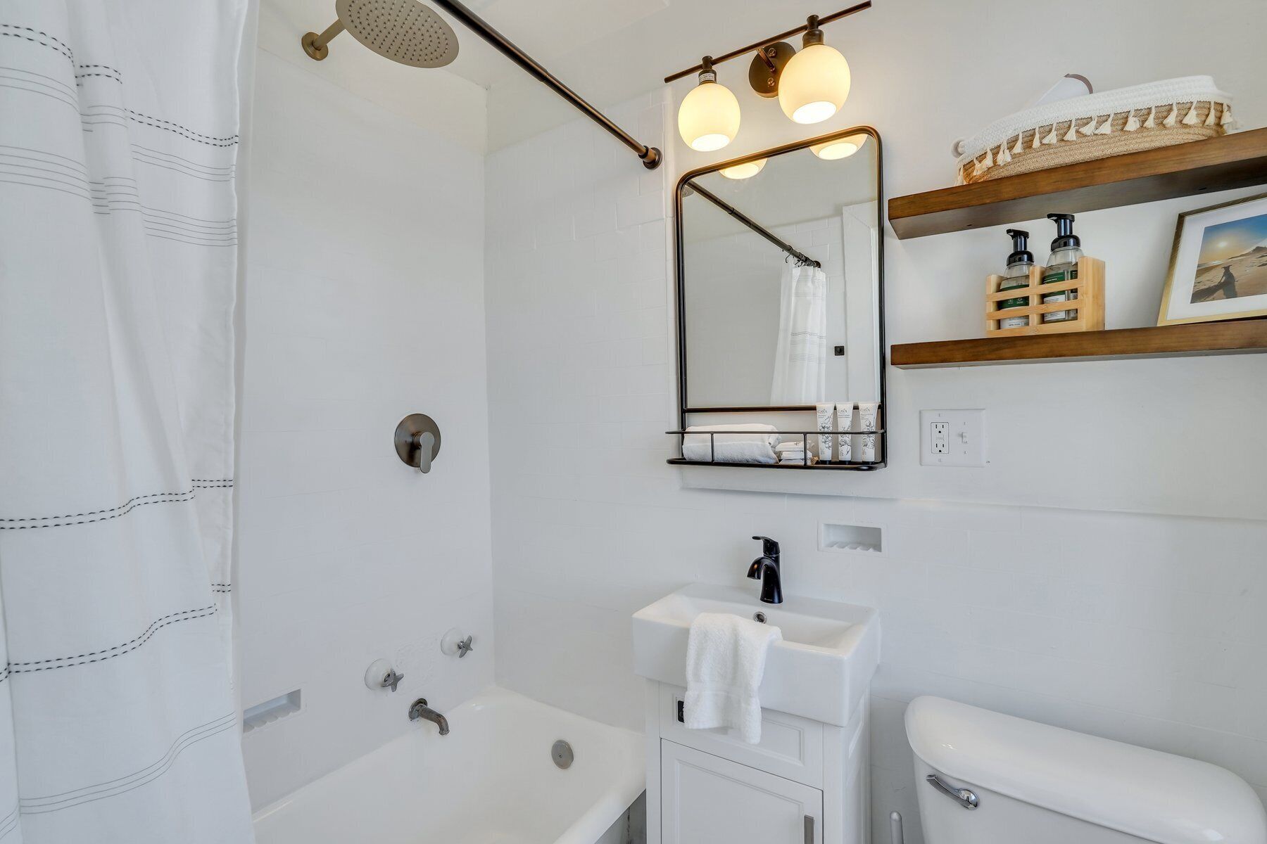 Ein modernes Badezimmer mit Dusche, Waschbecken, Toilette und Spiegel. Drei Lampen leuchten über dem Spiegel, und ein Handtuchhalter mit Handtüchern ist vorhanden.