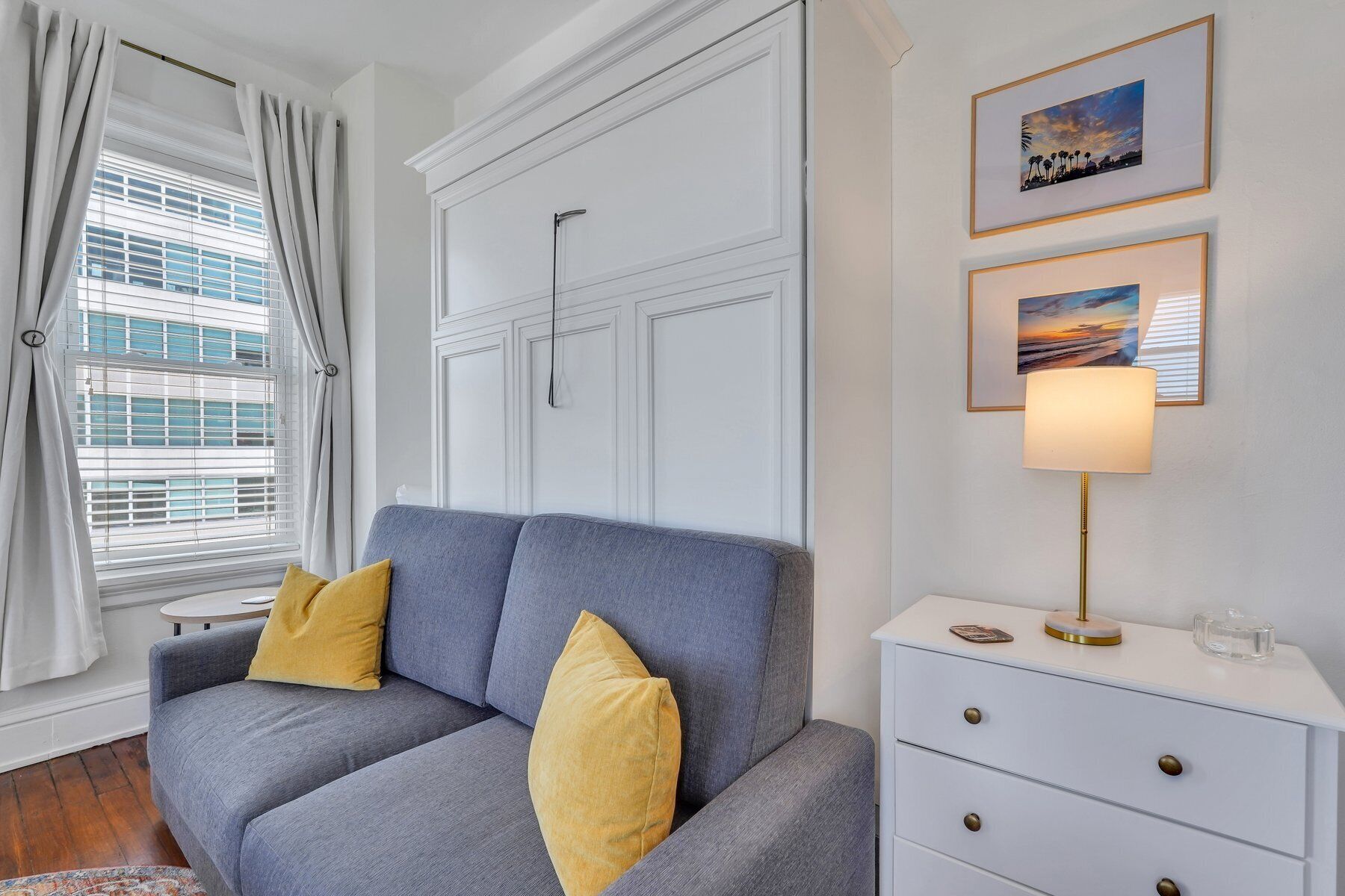 A modern living room with a grey sofa, yellow cushions, a white dresser with drawers, and framed artwork on the wall.