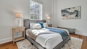 A bedroom with a large bed, two bedside tables, a window, and a painting on the wall. - Unbeatable Location in Heart of Historic Savannah by Lucky Savannah (Savannah)