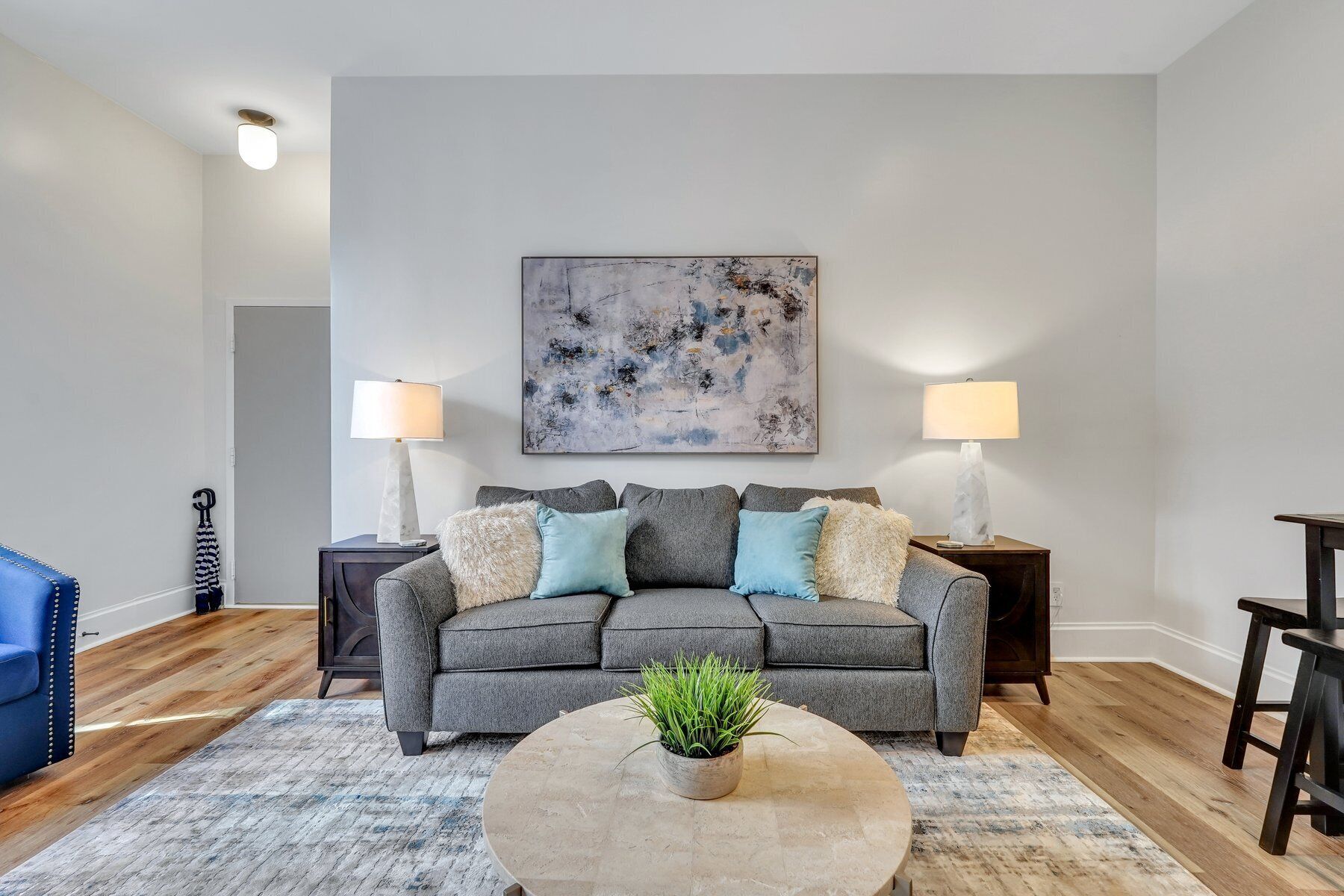 Un salon avec un canapé gris, deux tables d’appoint avec des lampes, une table basse avec une plante et un grand tableau abstrait accroché au mur.