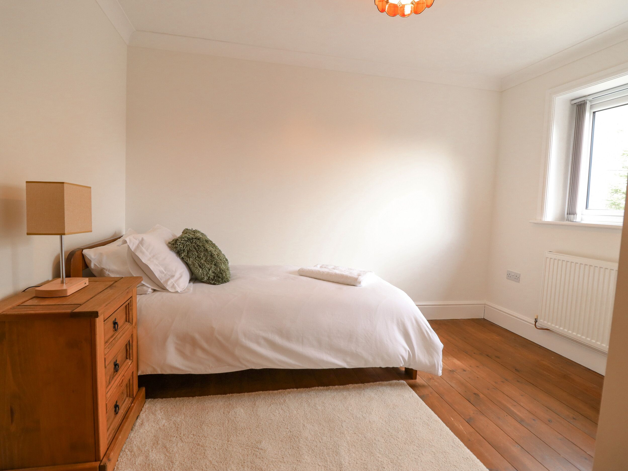 A bedroom with a bed, a wooden nightstand, a lamp, a radiator, and a window.