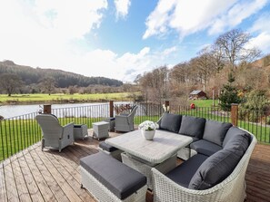 Una terraza con muebles de mimbre, una mesa y vistas a un lago y colinas.