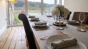 Mesa de comedor para cuatro personas con vista a un lago y un bosque a través de amplios ventanales.