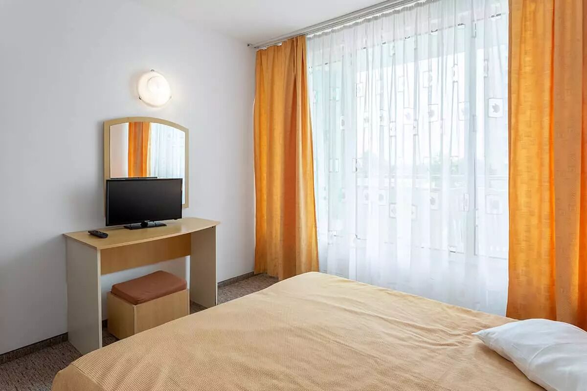 A hotel room with a bed, a desk with a TV, a chair, and a window with curtains.