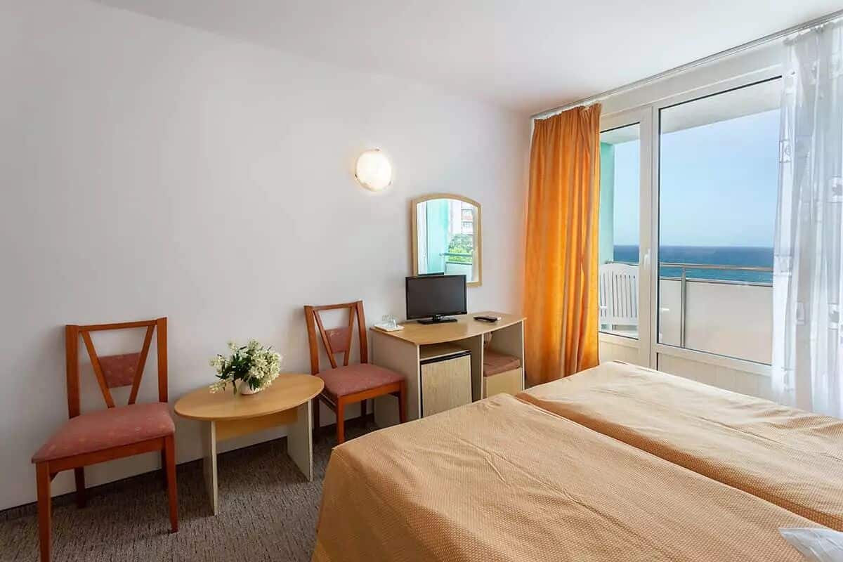 Una habitación de hotel con cama, dos sillas, un escritorio con televisor y un ventanal con cortinas.