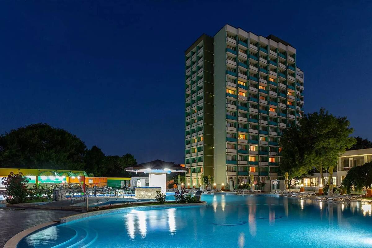 Un hotel de gran altura con piscina en la base, rodeado de árboles y un pequeño edificio con techo abovedado.