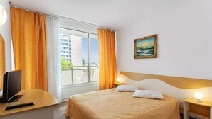 A hotel room with a bed, a desk with a TV and remote, a painting on the wall, and a balcony with curtains. - Hotel Balada (Saturn)