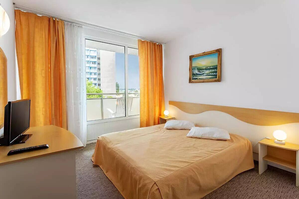 Una habitación de hotel con cama, escritorio con televisor y control remoto, un cuadro en la pared y un balcón con cortinas.