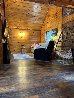 A cozy cabin interior with a wooden ceiling, a sofa, a hanging chair, and a fireplace.