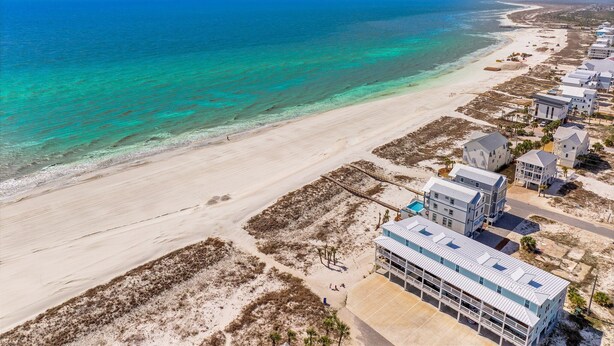 An aerial view of a coastal area with a beach, clear turquoise waters, and a row of modern beachfront villas with balconies.
