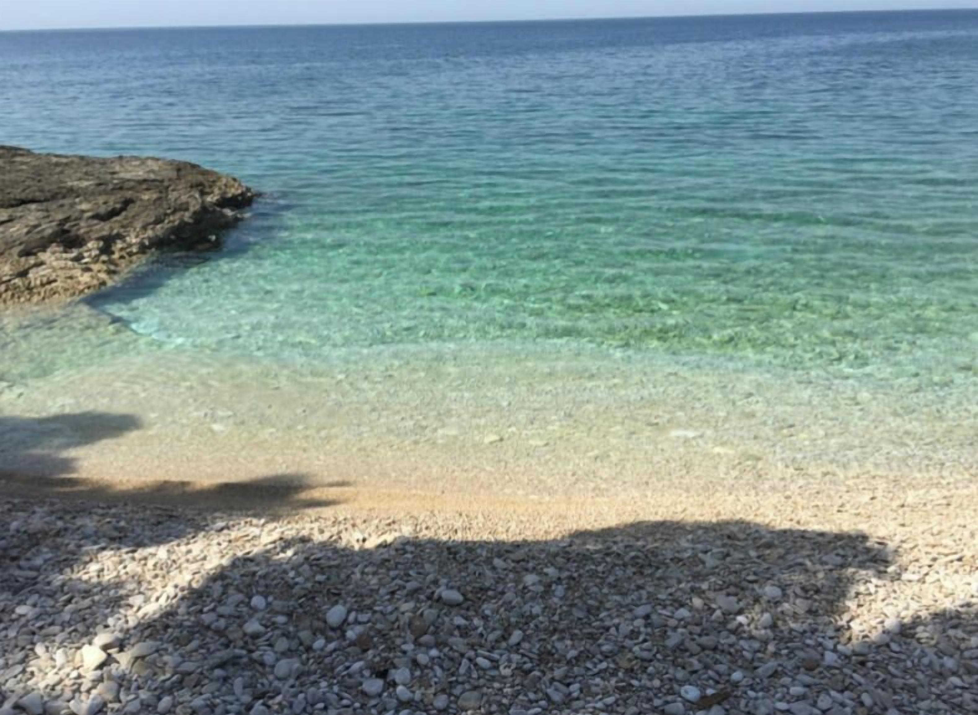 A pebble beach leading to clear turquoise water, with a rocky outcrop on the left.